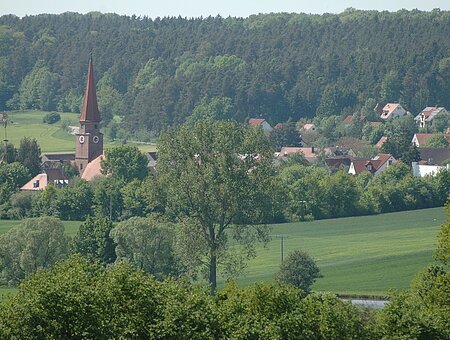 Eine grüne Landschaft mit Wiese und Bäumen. Darin ein roter Kirchturm und vereinzelte Häuser eines Dorfs